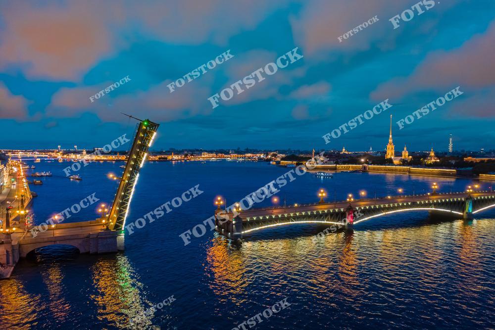На переднем плане изображен разведенный Троицкий мост, освещенный яркими огнями. Мост перекинут через реку Неву и соединяет центр Санкт-Петербурга с Петроградской стороной. На заднем плане видна Петропавловская крепость с ее высокими шпилями, освещенными в ночное время. Воды Невы отражают огни города, создавая эффектную ночную панораму.